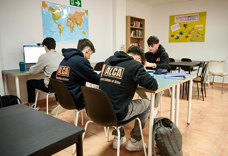 4 students in a bright, modern classroom showing desks, a computer and other study items