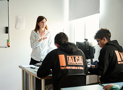 A teacher standing in front of 2 students in a white classroom