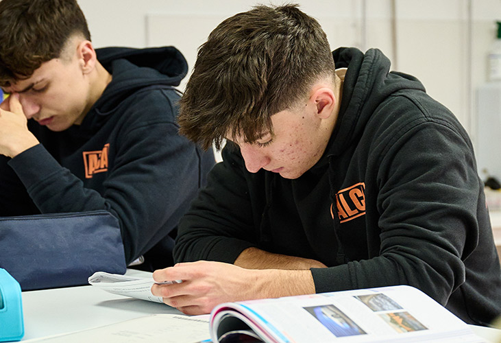 2 boys in black hoodies reading study material at a white table in a brightly lit room