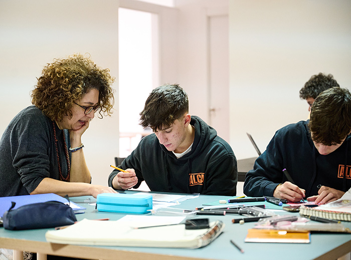 A female teaches with 3 male students sitting at desks in a classroom